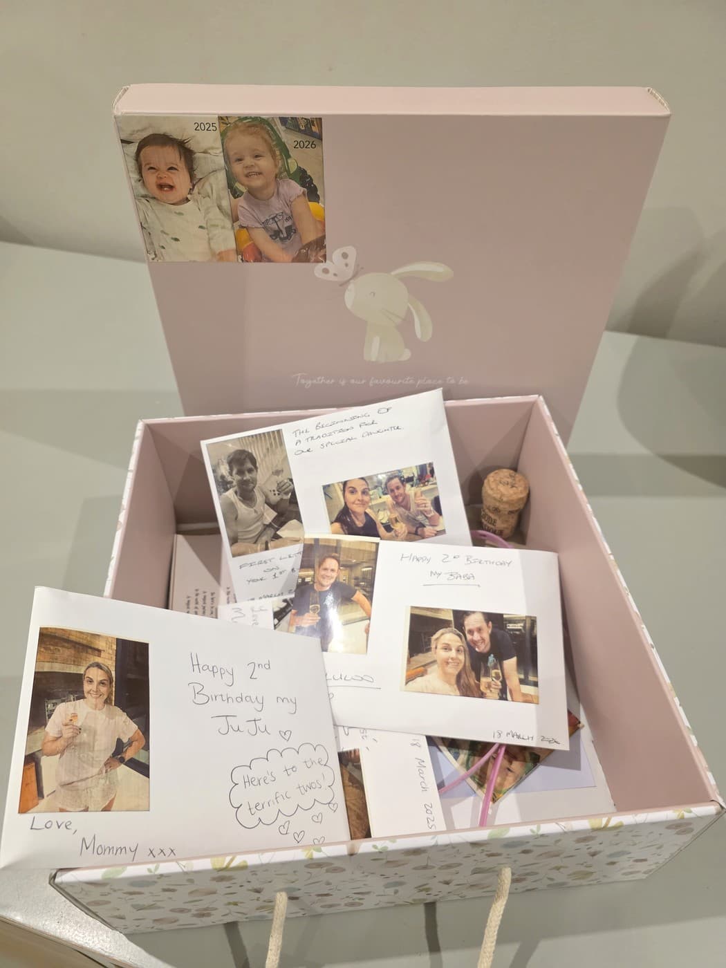 A pink keepsake box open on a table, containing handwritten letters, a champagne cork, and photographs of Julia on the inside of the lid.